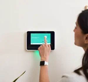 Woman interacting with a wall-mounted smart home alarm touchscreen panel, managing wireless security settings and monitoring features in a modern home environment.