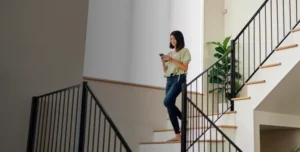 Woman walking down stairs while checking her smartphone, using a smart home security app to manage wireless alarm settings and monitor her alarm system remotely.