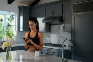 Woman standing in a modern kitchen using the Vivint smart home security mobile app to check system status, demonstrating convenient wireless alarm system control.